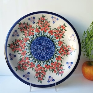 May include: A white ceramic plate with a blue rim and a floral design. The plate features a central blue pattern surrounded by red and pink flowers and blue accents. The plate is displayed on a clear stand.