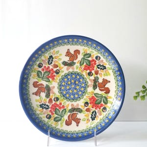May include: A round ceramic plate with a blue rim and a central design of a blue and yellow floral pattern. The plate features a repeating pattern of squirrels, berries, and leaves in shades of red, green, and brown.