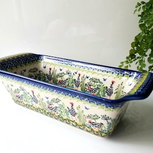 May include: Rectangular ceramic baking dish with a blue rim and handles. The dish features a hand-painted design of chickens, flowers, and foliage in shades of blue, green, and red. The dish is approximately 12 inches long.