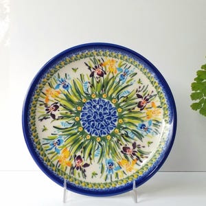 May include: A round ceramic plate with a blue rim and a floral design. The plate features a central blue floral pattern surrounded by green leaves and colorful flowers in yellow, blue, and purple. The plate is supported by a clear stand.