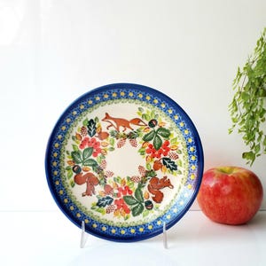 May include: A decorative ceramic plate with a blue rim and a detailed forest scene. The plate features a jumping fox, squirrels, and a wreath of leaves and berries. A red apple sits beside the plate.