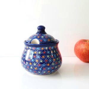 May include: A blue ceramic sugar bowl with a lid, adorned with a colorful polka dot pattern. The bowl has a small opening for a spoon. A red apple sits to the right of the bowl.