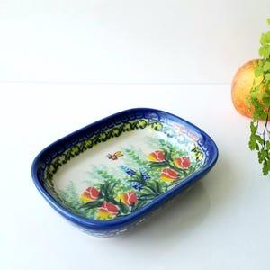 May include: Rectangular ceramic dish with a blue rim and floral design. The interior features red and yellow tulips, a bee, and other flowers. The dish is on a white surface.