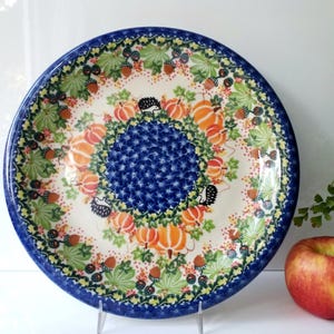 May include: A round ceramic plate with a blue rim and a central blue floral design. The plate features a decorative pattern of pumpkins, leaves, and hedgehogs. An apple rests beside the plate.