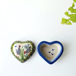 May include: A heart-shaped ceramic trinket box with a lid. The lid features a painted design of two black and white polka-dot chickens among green grass and small flowers. The base is cream-colored with a blue rim and three small blue birds inside.