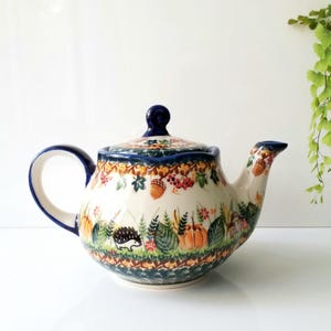 May include: A ceramic teapot featuring a cream-colored base with a hand-painted autumn-themed pattern. The design includes small hedgehogs, orange pumpkins, brown acorns, green leaves, and red berries. It has a dark blue handle, lid, and rim.