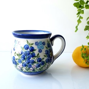 May include: A white ceramic mug with a blue rim and handle. The mug is decorated with a blue and green floral pattern featuring blueberries.