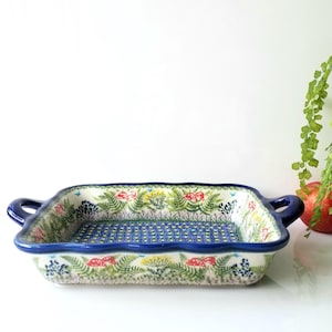 May include: A rectangular ceramic baking dish with two handles, featuring a blue rim and a decorative pattern of green ferns, yellow flowers, and red mushrooms on the sides, with a blue and yellow geometric pattern on the interior base.