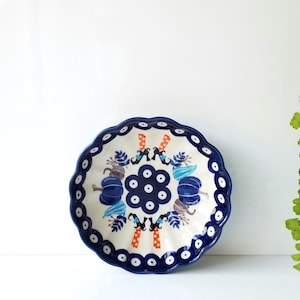 May include: A round ceramic bowl with a scalloped edge, featuring a cream-colored interior decorated with blue pumpkins, orange and white polka-dot witch legs, and dark blue floral accents. The rim is painted with a dark blue and white eye pattern.