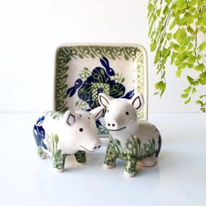 May include: A ceramic salt and pepper pig set with a matching square dish. The pigs are white with blue and green floral designs. The dish has a similar pattern with a rabbit motif. A charming kitchen decor item.