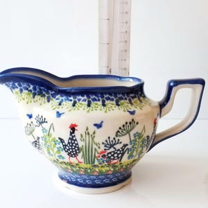 May include: A white ceramic gravy boat with a blue and green floral pattern featuring chickens. The handle is on the right side of the boat.