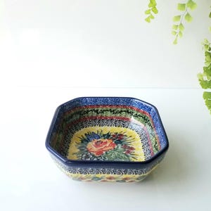 May include: An octagonal ceramic bowl featuring a vibrant floral pattern with a central orange rose, surrounded by green leaves and small blue flowers on a yellow background, finished with a dark blue rim and intricate decorative border.