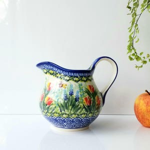 May include: A ceramic pitcher with a blue rim and handle, decorated with a floral pattern of red tulips, blue flowers, and green leaves. The pitcher has a white interior and sits on a white surface. An orange apple and greenery are in the background.
