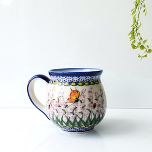 May include: A ceramic mug with a blue handle and rim, featuring a floral and butterfly design. The mug has a white base with a pattern of pink flowers, green grass, and a yellow and orange butterfly. The mug is approximately 4 inches tall.