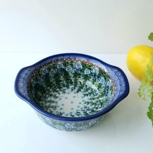 May include: A ceramic serving bowl with a blue rim and handles. The bowl features a floral pattern in shades of blue and green. A yellow lemon and green foliage are in the background.