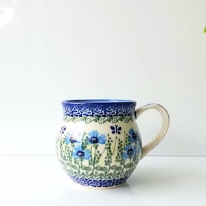 May include: A round ceramic mug featuring a hand-painted pattern of blue cornflowers, green stems, and small dark blue butterflies. The mug has a dark blue decorative border at the top and bottom, a curved handle, and a wide, bulbous body shape.