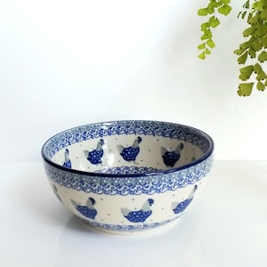 May include: A round ceramic bowl featuring a cream-colored base with a repeating pattern of blue chickens. The design includes intricate blue floral scrollwork and small dot accents along the rim and midsection, creating a detailed decorative motif.