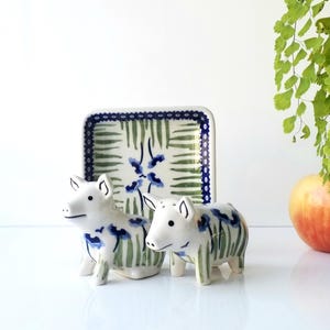 May include: Two white ceramic pig figurines with blue and green floral accents stand before a matching rectangular dish. The dish has a blue border and a green and blue floral pattern. An apple and greenery are in the background.