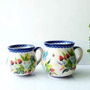 May include: Two ceramic mugs with a rounded shape, featuring a hand-painted pattern of red strawberries, green leaves, small blue flowers, and a bright blue butterfly. The rims are decorated with a repeating blue scalloped border design.