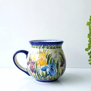 May include: A round ceramic mug featuring a hand-painted floral pattern with yellow, blue, and purple irises on a cream background. The mug has a dark blue rim, a matching dark blue handle, and a decorative border pattern along the top and base.