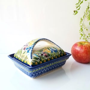 May include: A ceramic butter dish with a lid and handle, featuring a floral design in blue, yellow, and purple. The dish has a blue border and sits next to a red apple. The butter dish is a kitchenware item.