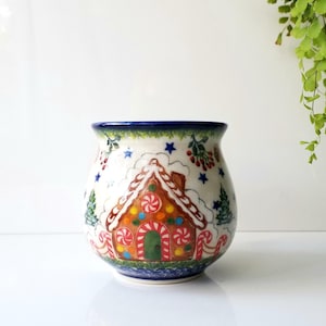 May include: A ceramic jar with a festive gingerbread house design. The jar features a white base with a colorful gingerbread house, candy canes, and Christmas tree illustrations. The rim and base have blue accents.
