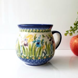 May include: A ceramic mug with a floral design featuring yellow, blue, and purple irises on a white background. The mug has a dark blue rim and handle. An apple is next to the mug.