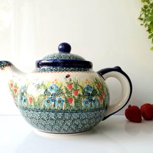 May include: A ceramic teapot with a lid, featuring a floral design with blue flowers and red strawberries. The teapot has a white background with a blue rim and handle. Three fresh strawberries are next to the teapot.