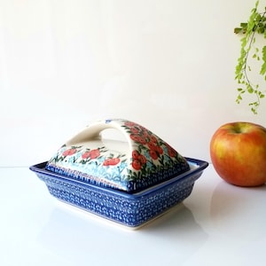 May include: A rectangular ceramic butter dish with a lid and handle, decorated with red floral patterns on a white background. The base is blue with a dotted design. An apple and greenery are in the background.