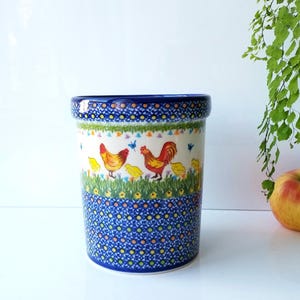 May include: A cylindrical ceramic utensil holder with a blue rim and base, featuring a hand-painted design of chickens, roosters, and chicks in a grassy field. The holder is approximately 6 inches tall.