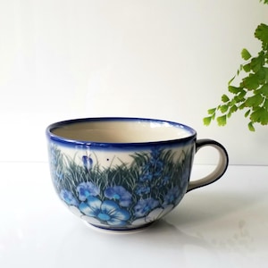 May include: A ceramic teacup with a blue floral design. The cup has a white interior and a blue rim. The exterior features a pattern of blue flowers and green foliage. The handle is black. The cup is approximately 4 inches in diameter.