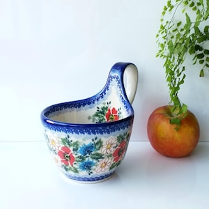 May include: A ceramic serving bowl with a handle, featuring a floral pattern in red, blue, and white. The bowl has a dark blue rim and handle. An apple and greenery are in the background.