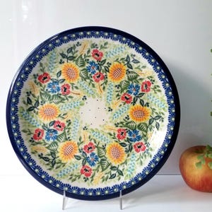 May include: A round ceramic plate with a dark blue rim and a floral design. The plate features yellow sunflowers, red and blue flowers, and green leaves. The plate is on a clear stand.