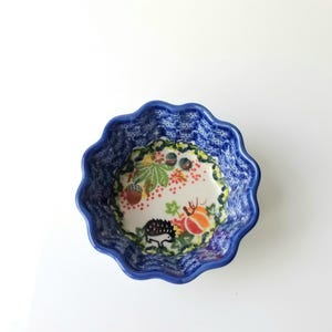 May include: A scalloped ceramic bowl featuring a deep blue exterior with a textured pattern. The interior base displays a cream-colored surface decorated with a colorful illustration of a small hedgehog, an orange pumpkin, and brown acorns.