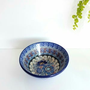 May include: A round ceramic bowl featuring a detailed floral pattern in shades of blue, white, and brown. The interior and exterior rim are decorated with a repeating geometric border, while the center displays larger blue and brown flower motifs.