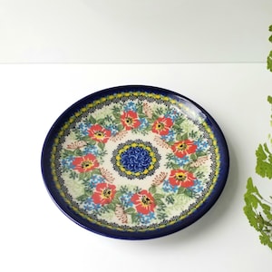 May include: A round ceramic plate with a dark blue rim and a floral pattern. The plate features red flowers with yellow centers, blue accents, and a central blue floral design. The plate is on a white surface.