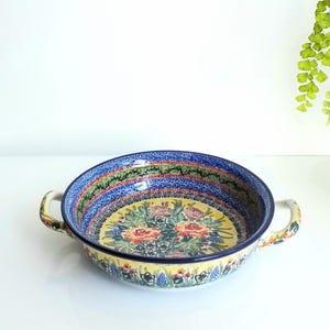 May include: A round ceramic baking dish with two side handles, featuring a vibrant floral pattern with yellow, red, and blue colors. The interior base displays a bouquet of roses, while the rim is decorated with intricate blue and green patterns.