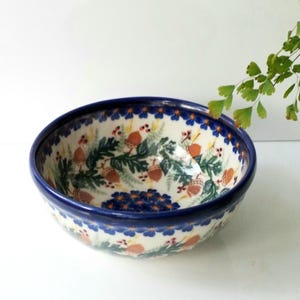 May include: A ceramic bowl with a blue rim and a detailed interior design. The bowl features a pattern of acorns, green leaves, and blue flowers on a cream background. The bowl is a decorative piece.