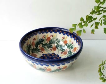 May include: A ceramic bowl with a blue rim and a detailed interior design. The bowl features a pattern of acorns, green leaves, and blue flowers on a cream background. The bowl is a decorative piece.