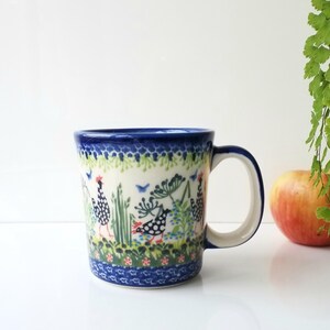 May include: A ceramic mug with a blue rim and handle, featuring a hand-painted design of chickens, flowers, and foliage. The mug has a white background with a blue and green border. An apple is next to the mug.