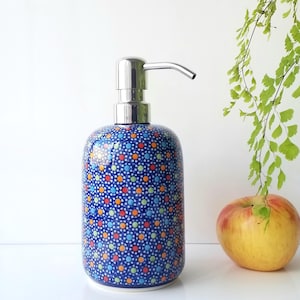 May include: A blue ceramic soap dispenser with a stainless steel pump. The dispenser is decorated with a pattern of small, colorful dots. An apple and green foliage are in the background.