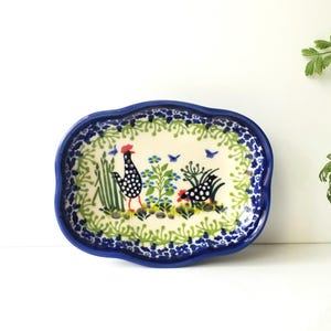 May include: A ceramic serving dish with a scalloped edge, featuring a hand-painted design. The dish has a blue border and a cream-colored interior with a scene of two chickens, plants, and butterflies. The dish measures approximately 7 inches long and 5 inches wide.