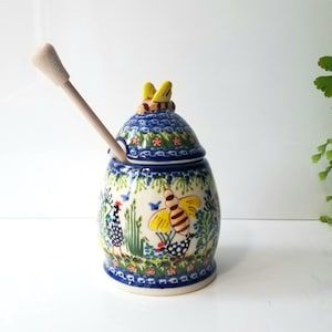 May include: A ceramic honey pot with a wooden dipper. The pot is decorated with a blue floral pattern and a yellow bee design. The lid has a bee-shaped handle. The honey pot is approximately 5 inches tall.