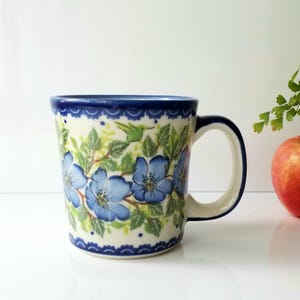 May include: A ceramic mug with a blue floral design. The mug features a white base with blue flowers and green leaves. The rim and handle are blue. An apple and greenery are in the background.