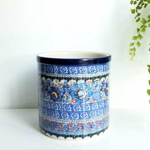 May include: A cylindrical ceramic utensil holder featuring a dark blue rim and a multi-layered pattern of blue, white, and red floral and geometric motifs. The exterior surface is decorated with horizontal bands of intricate designs and small flowers.