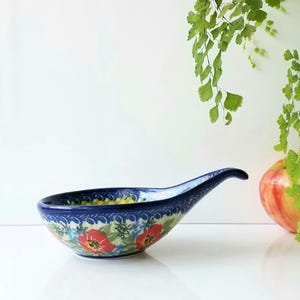 May include: A blue ceramic spoon rest with a floral design featuring red flowers and green leaves. The spoon rest has a curved handle and a decorative rim. A red and yellow apple and green foliage are in the background.