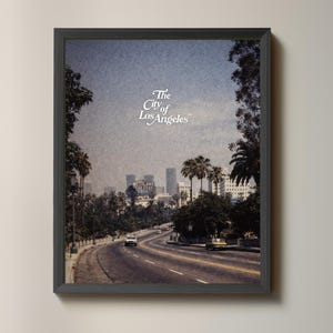 May include: Framed print featuring a vintage view of Los Angeles, California. The image shows a road leading towards the city skyline, with palm trees lining the sides. The text "The City of Los Angeles" is displayed in white script at the top.