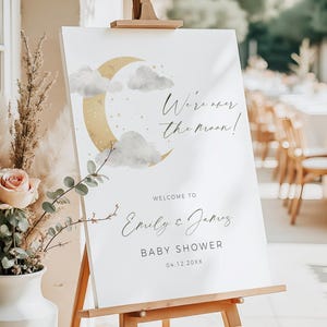 May include: A white baby shower welcome sign on a wooden easel. The sign features a gold crescent moon, clouds, and the text "We're over the moon!" and "Welcome to Emily & James". The date is 04.12.20XX.