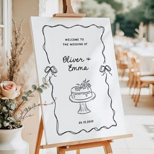 May include: A white wedding welcome sign on a wooden easel. The sign features black script that reads "Welcome to the Wedding of Oliver & Emma" with a cake illustration and the date 06.12.20XX.