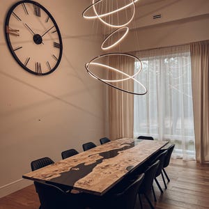 May include: A large dining table with a live edge epoxy resin top and black chairs. The table is made of wood with a natural grain pattern. A modern light fixture with multiple circular rings hangs above the table.